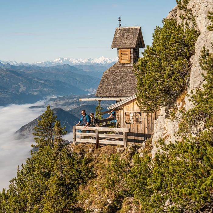 Wanderer am Aussichtspunkt an einer Holzhütte auf einem Berg mit Blick auf Täler und Berge.