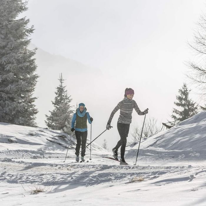 Zwei Frauen beim Skilanglauf auf einem schneebedeckten Pfad, umgeben von beschneiten Bäumen und nebligem Hintergrund.