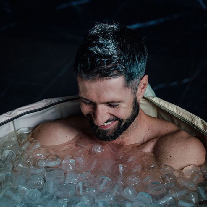 Man with a beard, smiling, in an ice bath with ice cubes, looking down.