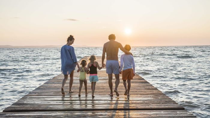 Famiglia di quattro persone che cammina insieme su una passerella di legno verso il mare al tramonto.