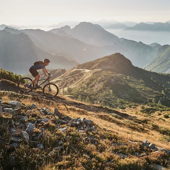 A cyclist riding a mountain bike on a rugged trail with scenic mountain views at Falkensteiner Hotels & Residences