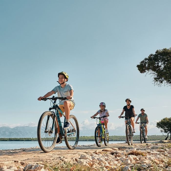 A family cycling along a stony beachside path with a clear sky in the background at Falkensteiner Hotels & Residences