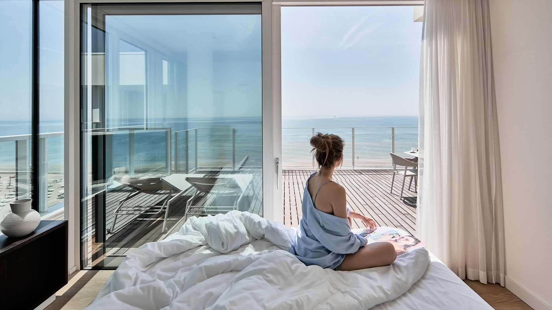 Person sitting on a bed facing the sea view from a beachfront room with open terrace doors at Falkensteiner Hotel & Spa Jesolo