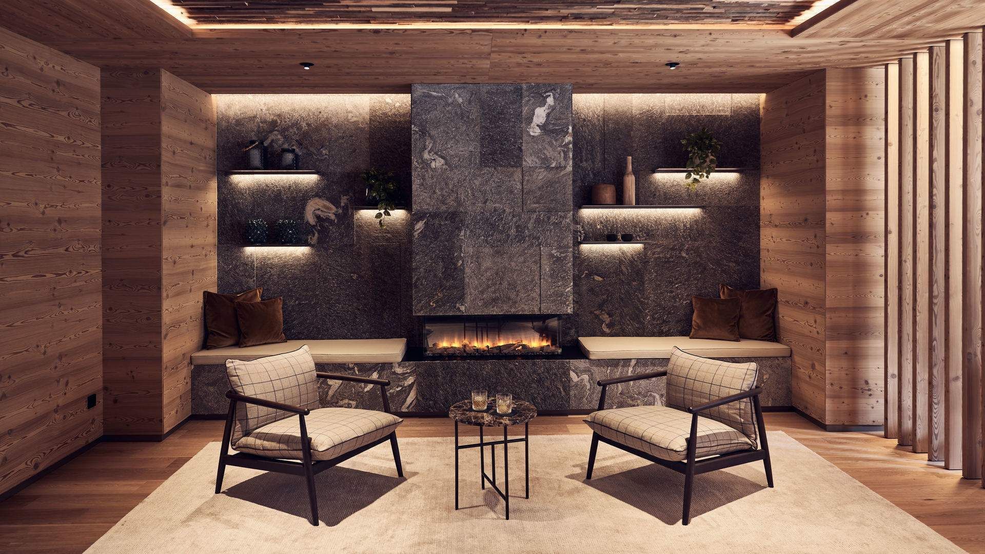 Cozy living space featuring a stone fireplace, two plaid chairs, and a coffee table at Falkensteiner Hotel Kronplatz