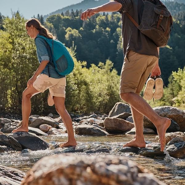 Hikers crossing stream on stepping stones with a forest backdrop near Falkensteiner Hotel Kronplatz