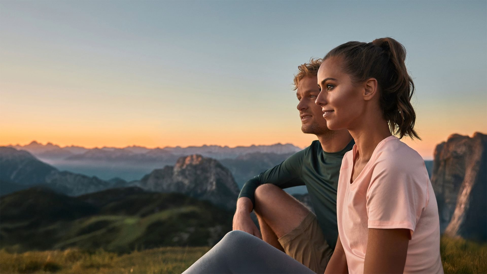 A man and a woman sit on a hill at sunset, overlooking mountains.