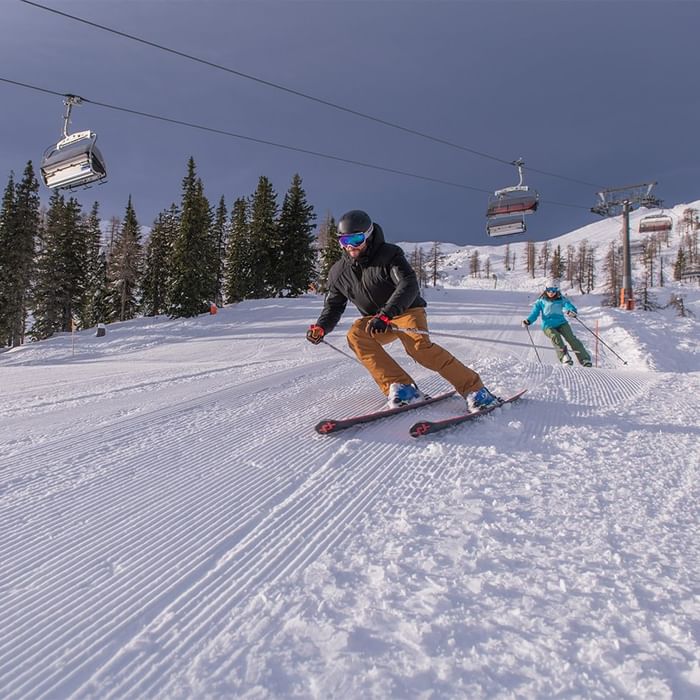 Zwei Skifahrer navigieren eine schneebedeckte Piste mit Skiliften im Hintergrund.