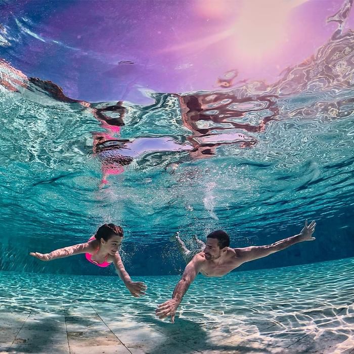 A man and a woman are swimming underwater in a pool, reaching out towards each other.