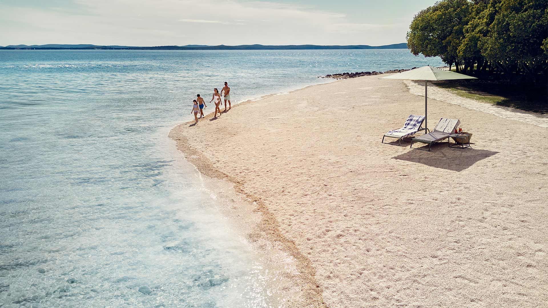 Obitelj uživa na privatnoj plaži s ležaljkama i suncobranom, okružena morom i šumom.