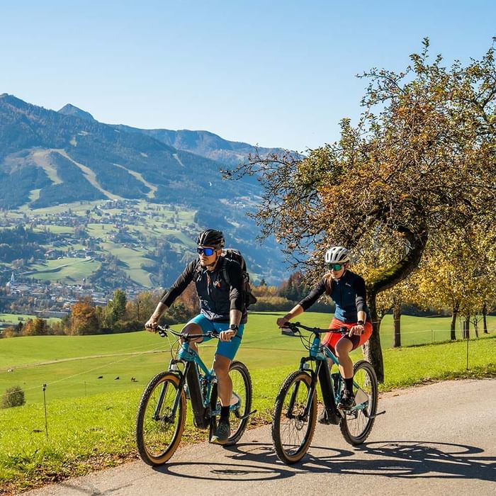 Zwei Radfahrer fahren auf einer Straße in ländlicher Umgebung mit Bergpanorama im Hintergrund.