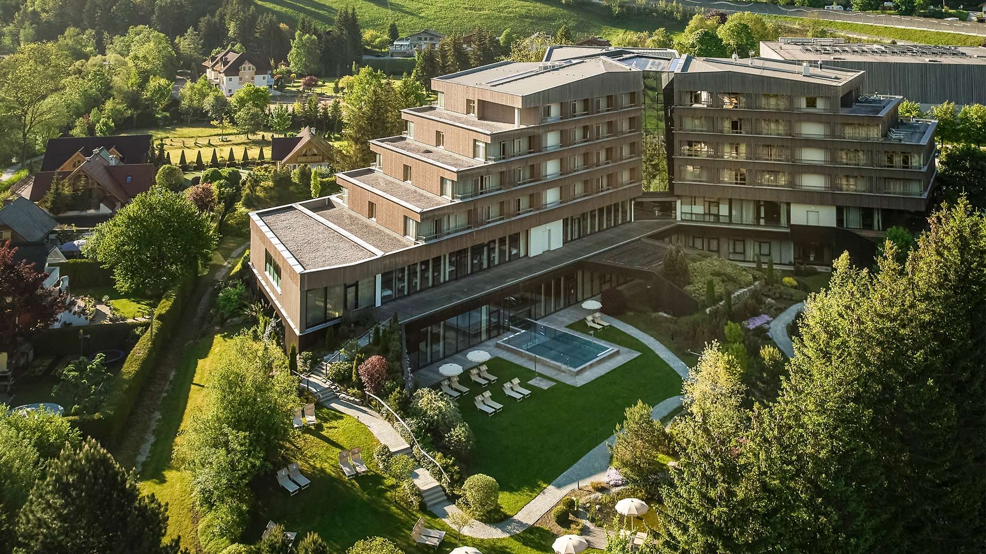 Luftaufnahme eines modernen Hotels mit Pool und Liegestühlen, umgeben von Bäumen und Gärten.