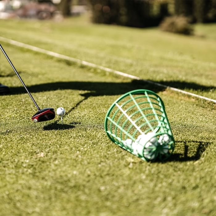 Golfschläger bereit zum Schlag, Ball auf Tee, grüner Korb mit Bällen, Grasfläche, verschwommener Hintergrund.