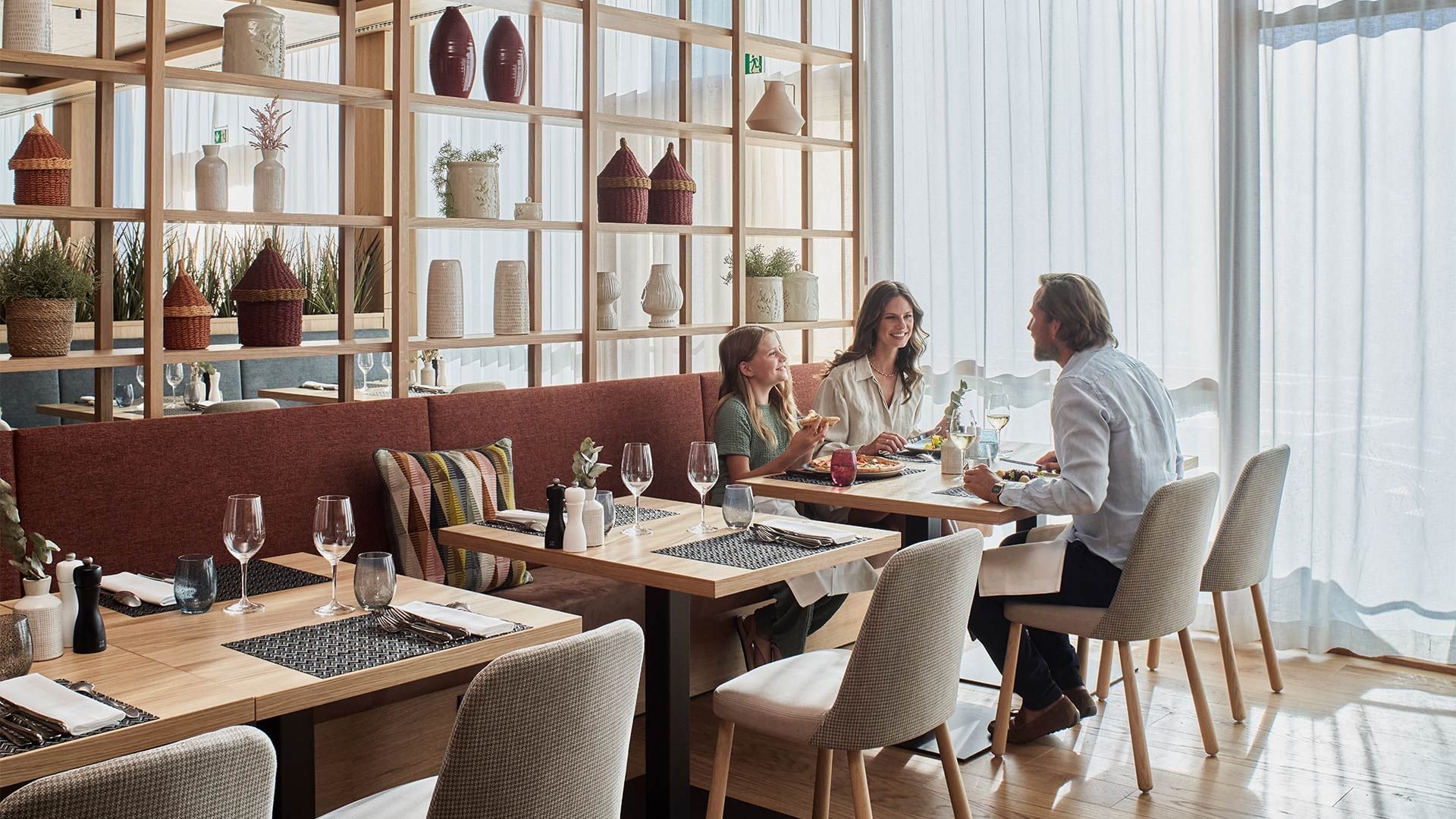 Three people dining in restaurant with decorative shelves and large windows at Falkensteiner Hotel and Spa Carinzia