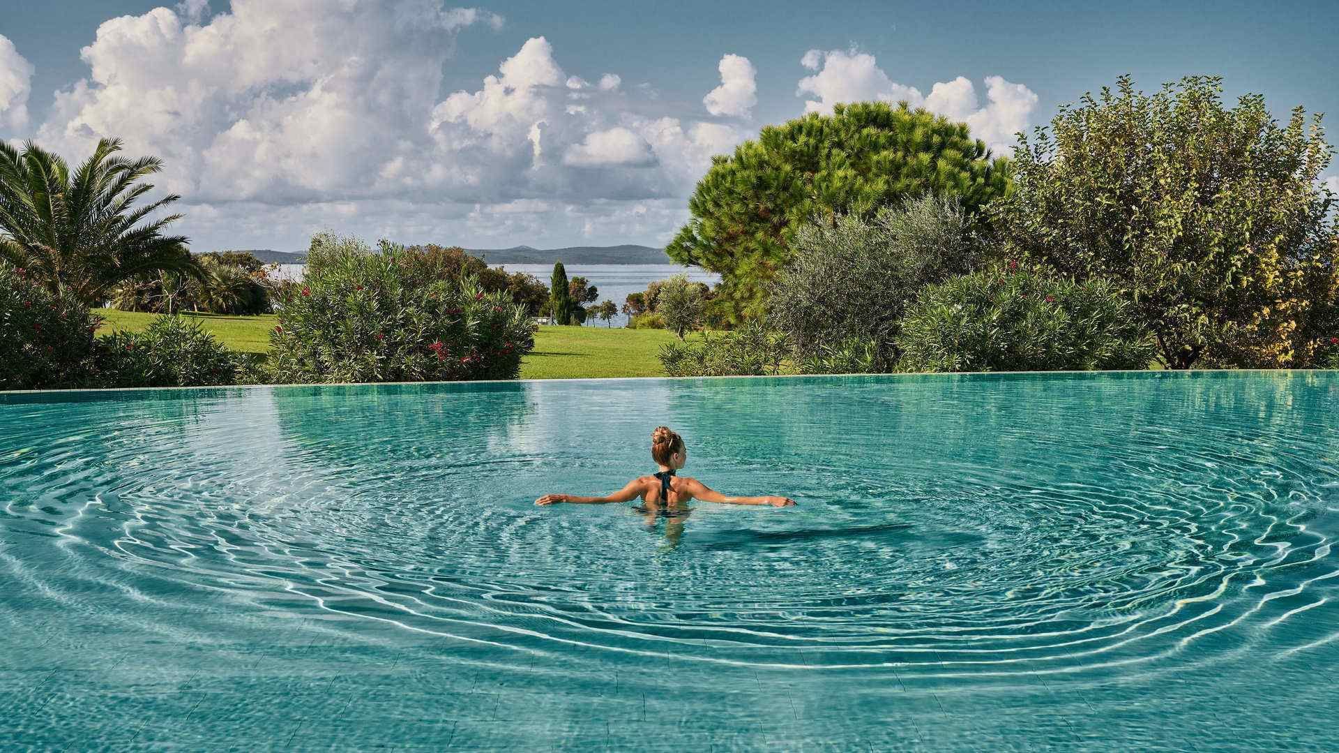 A woman swims in a large pool with trees and a cloudy sky in the background.