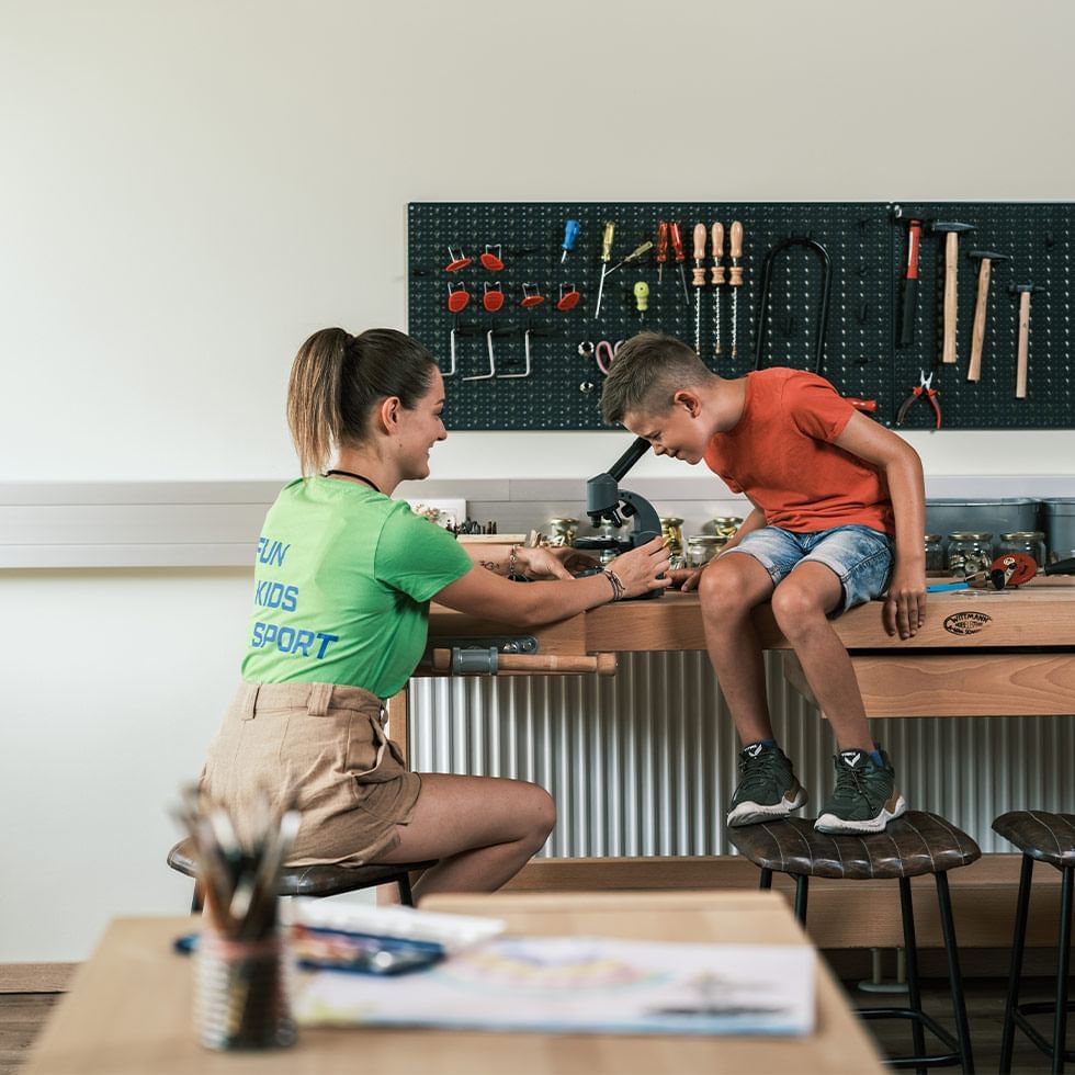 Adult assisting a child with a tool on a workshop table at Falkensteiner Club Funimation Katschberg