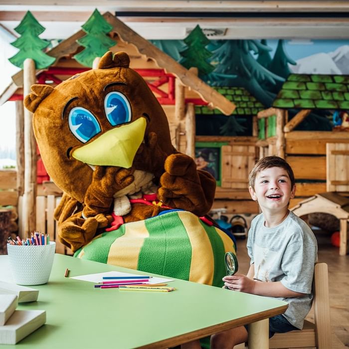Person in an owl costume sitting at a table with a kid & coloring materials at Falkensteiner Hotel Sonnenalpe