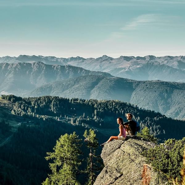 Two people sitting on a mountain edge overlooking a forested valley near Falkensteiner Hotel Montafon