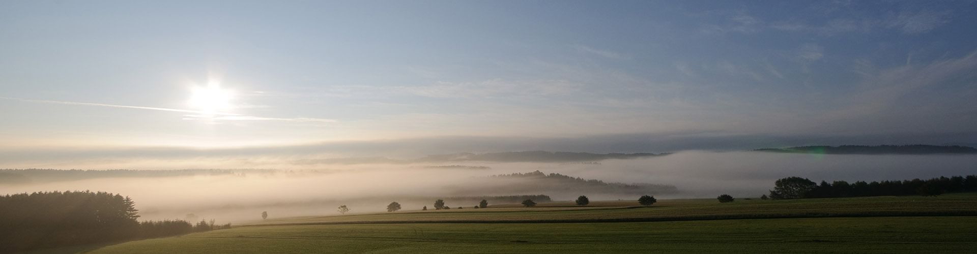 Panoramabild eines nebligen Tals mit Bäumen und Feldern, die Sonne strahlt hell am Himmel.