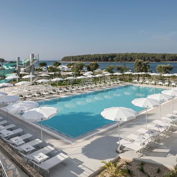 Luksuzni hotelski kompleks s velikim bazenom, ležaljkama, suncobranima i pogledom na more.