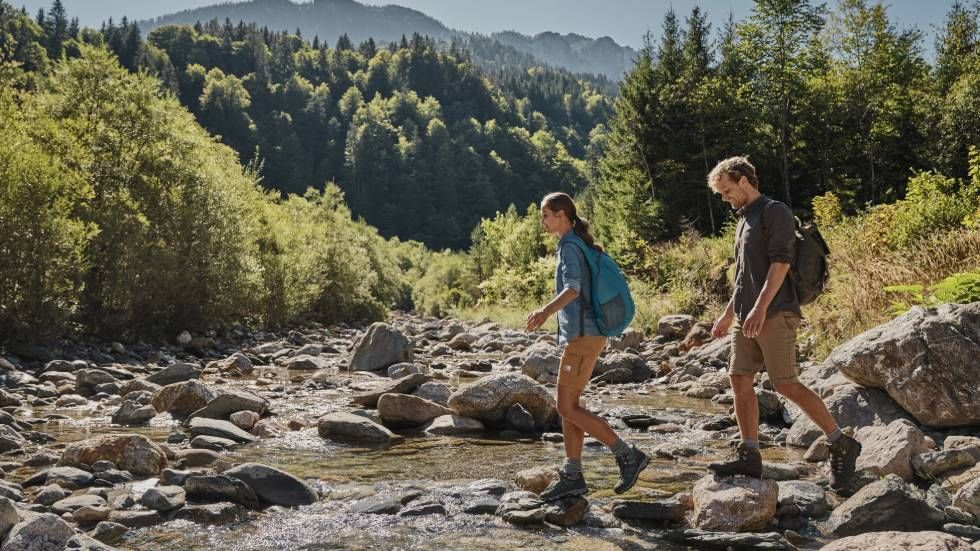 Two hikers crossing a rocky creek in a sunny forested landscape near Falkensteiner Hotels & Residences