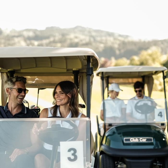 Two couples are riding golf carts, smiling, with a mountain view in the background.