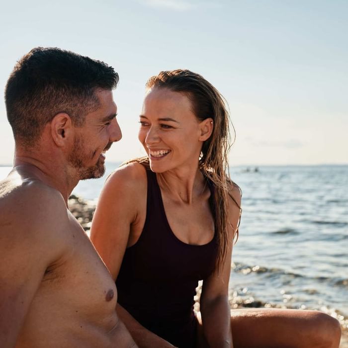 Couple sits together on the beach, gazing at the ocean under a bright sky at Falkensteiner Hotels & Residences