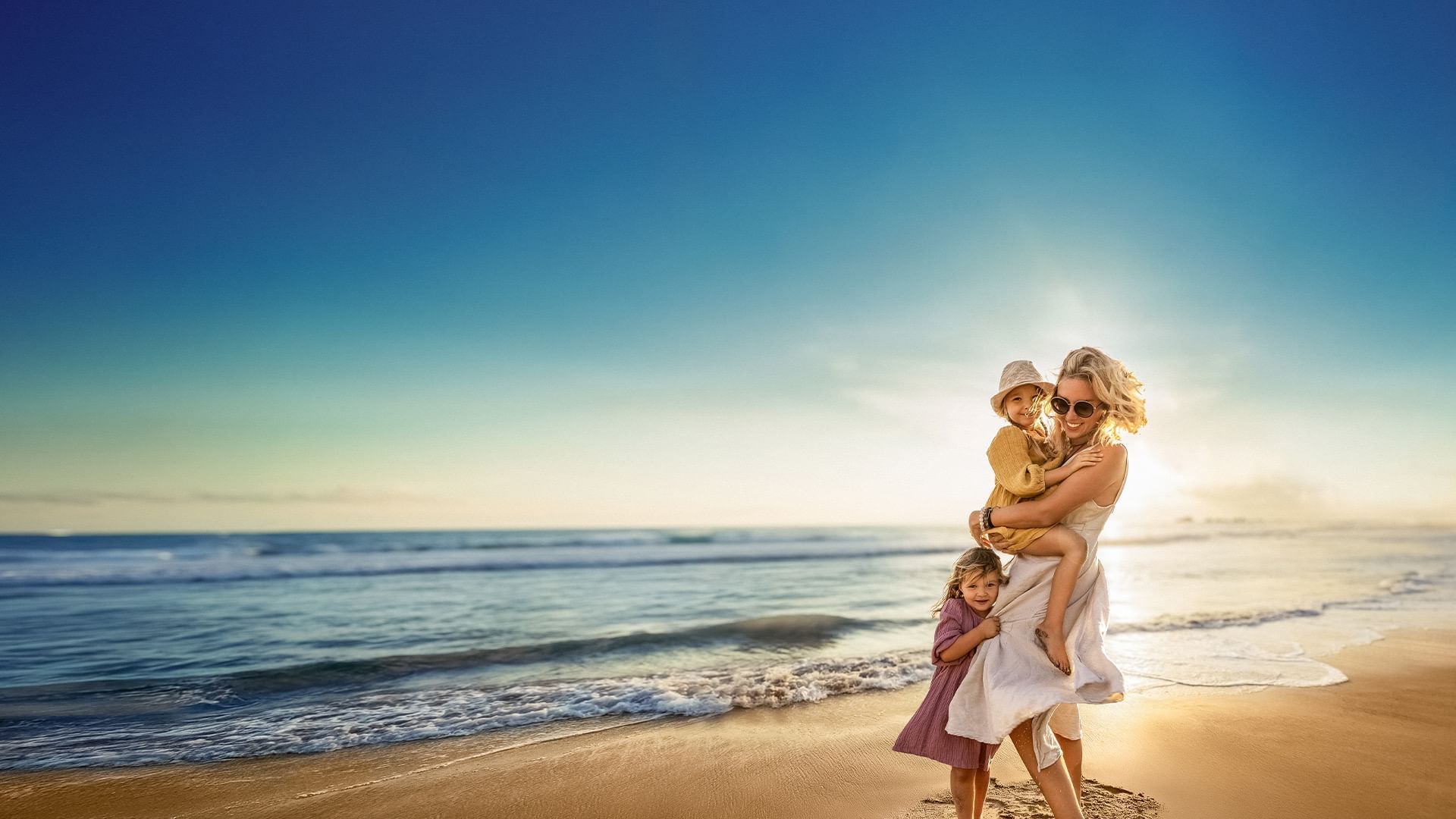 A mother on a beach holds one child on her hip while another stands nearby.