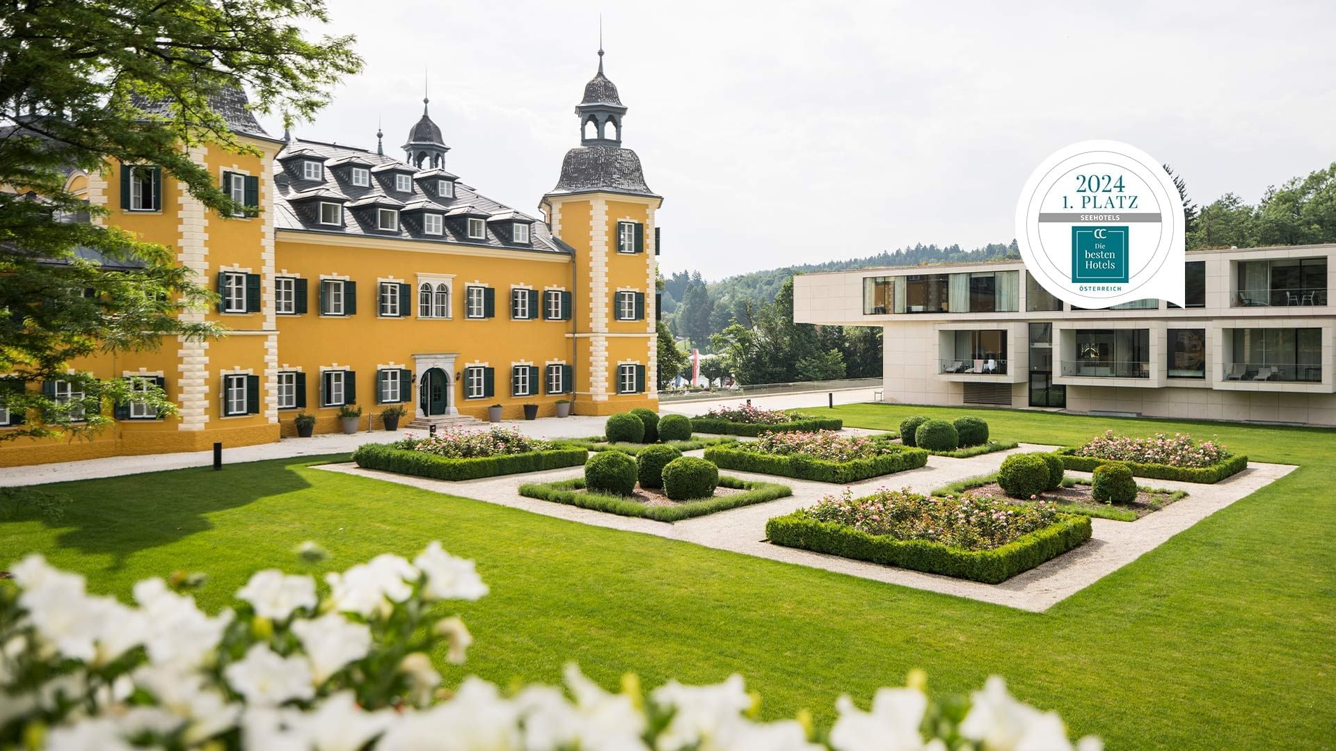 Gelbes Gebäude mit vielen Fenstern, Garten und weiße Blumen im Vordergrund, modernes Gebäude im Hintergrund.