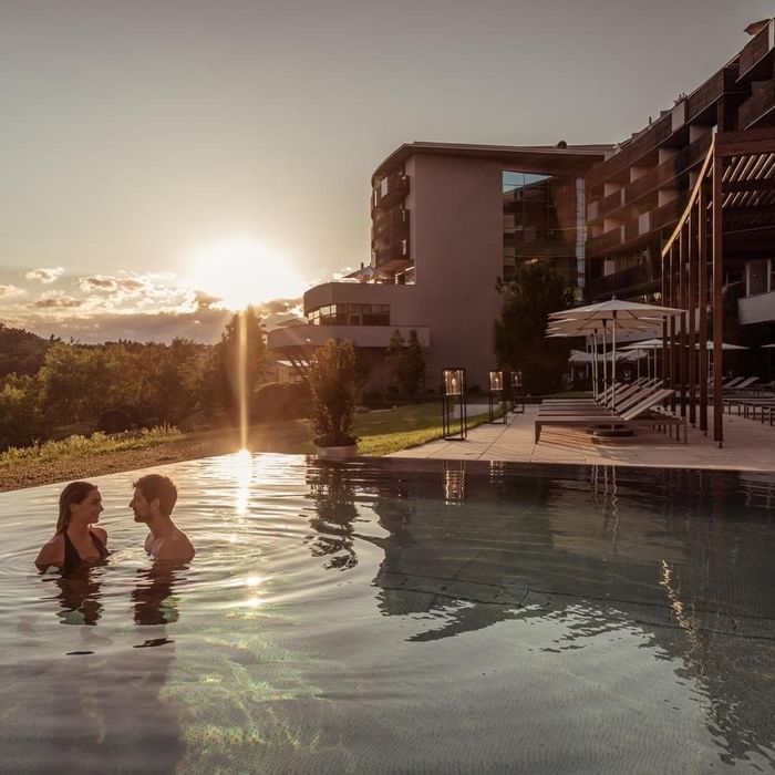 Due persone in una piscina all'aperto con un resort sullo sfondo al tramonto.